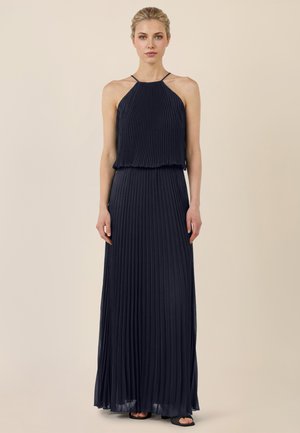 Kobieta w granatowej, plisowanej sukni maxi bez rękawów i z cienkimi ramiączkami, stojąca na tle jednolitego beżowego tła.