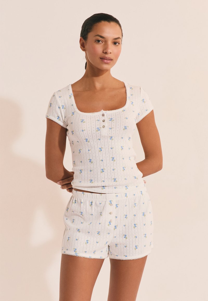 Femme debout, les bras derrière le dos, portant un haut court à manches courtes côtelé blanc à motifs floraux et un short assorti, sur fond beige.