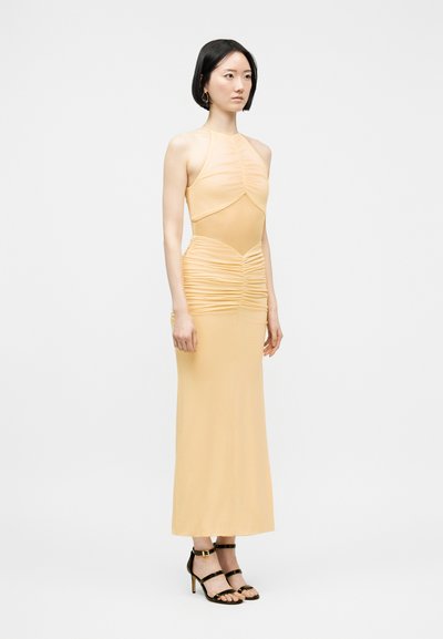 Γυναίκα με αμάνικο, ρούχο με πτυχές σε απαλό κίτρινο χρώμα και διαφανή μέση, φοράει μαύρα σανδάλια με λουράκια, στέκεται μπροστά σε απλό φωτεινό φόντο.