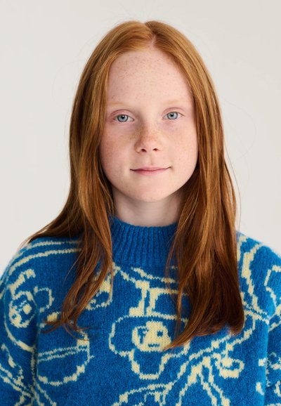 Fille rousse avec des taches de rousseur porte un pull bleu avec des motifs abstraits crème, regardant calmement devant un fond clair uni.