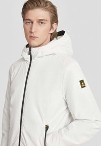 Giacca bianca con cappuccio, chiusura con zip frontale, zip nera e dettagli neri, texture liscia e un piccolo logo nero e giallo sulla manica sinistra.