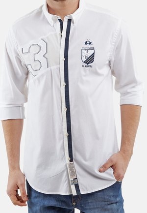 Mężczyzna w białej koszuli zapinanej na guziki z podwiniętymi rękawami, granatowymi lamówkami, naszywką z numerem 3 oraz logo Polo Club La Martina na piersi.