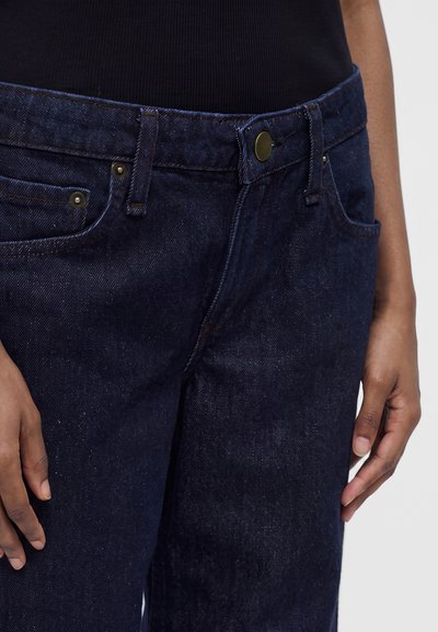 Gros plan d'une personne portant un jean en denim bleu foncé et un haut noir, les mains reposant sur les côtés sur un fond gris clair.