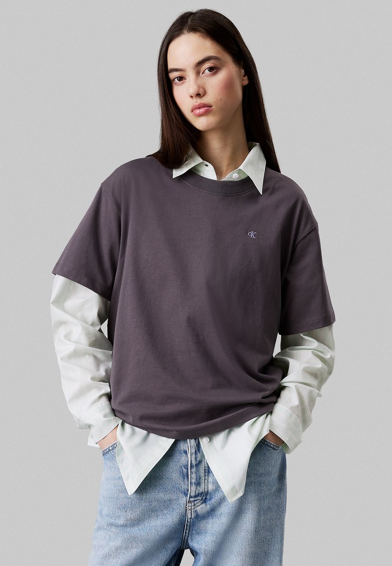 Camiseta de algodón púrpura superpuesta sobre una camisa de botones a rayas verde claro; corte relajado, mangas cortas y pequeño detalle de logo en el pecho.