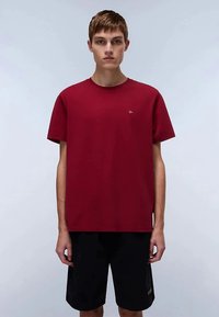 Jovem de pé, voltado para frente, usando uma t-shirt simples de manga curta vermelha e calções pretos, contra um fundo claro e liso.