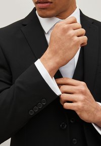 Black suit jacket with a white dress shirt. Visible black buttons on the cuff and a smooth texture. Hands adjusting the jacket.