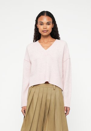 Suéter cropped rosa claro, com decote em V e punhos canelados, combinado com uma saia plissada em tom caqui. Com textura suave e um corte descontraído.