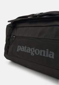 Black duffel bag with a textured surface, featuring a top zipper, side pocket, and "patagonia" logo in gray. Robust straps are attached.