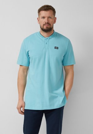 Mężczyzna z brodą ubrany w jasnoniebieską koszulkę polo z krótkim rękawem i dwoma guzikami oraz granatowe spodnie, stojący na jednolitym tle.