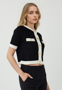 Cardigan corto nero con dettagli a coste bianche, dotato di bottoni dorati tondeggianti e due tasche sul petto. Indossato con pantaloni neri su misura.