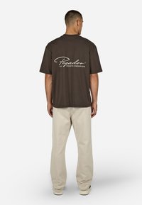 Camiseta de algodón marrón oscuro con el logo "Pegador" en blanco en la parte trasera, combinada con pantalones de corte relajado en beige claro.