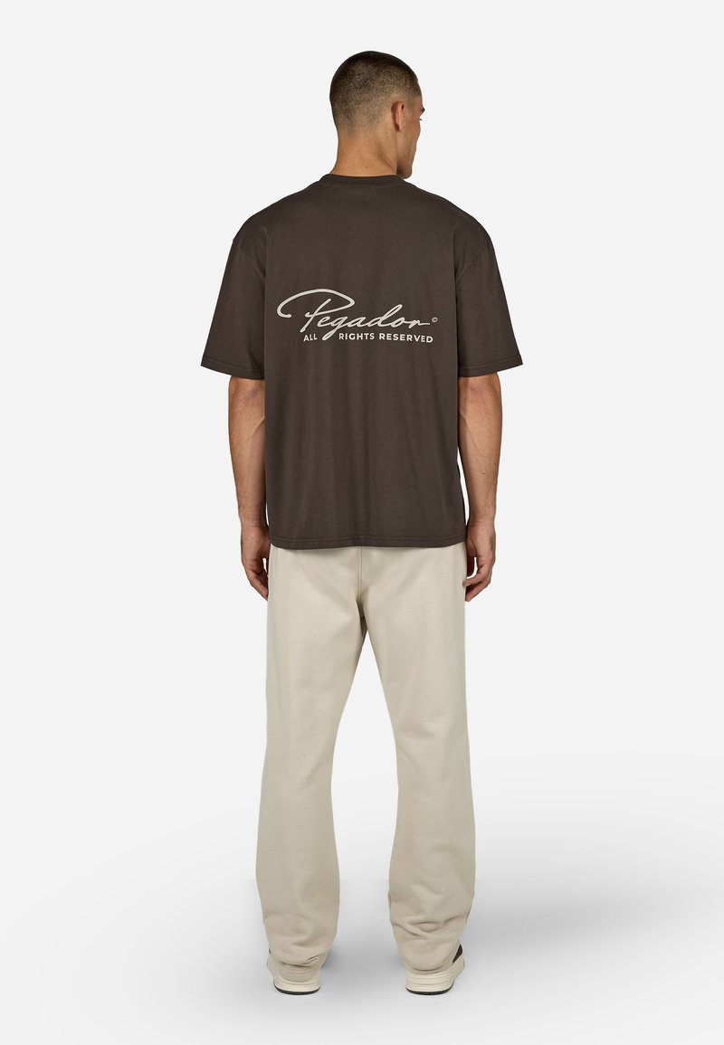 Camiseta de algodón marrón oscuro con el logo "Pegador" en blanco en la parte trasera, combinada con pantalones de corte relajado en beige claro.