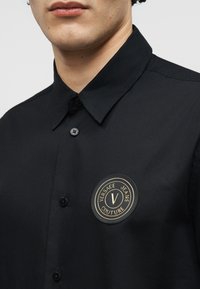 Άνδρας που φοράει μαύρο πουκάμισο με κουμπιά, με κυκλικό λογότυπο Versace Jeans Couture στο αριστερό στήθος.