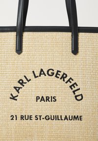 Šiaudinis krepšys su juodais odiniais rankenomis, su išsiuvinėtuku: "KARL LAGERFELD PARIS 21 RUE ST-GUILLAUME." Natūralus ir tekstūruotas paviršius.
