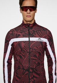 Röd och svart mönstrad zip-jacka med hög krage och vita sidostråk; inkluderar synlig logotyp på bröstet och strukturerad tygdesign.