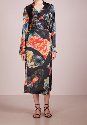 Kvinde iført en langærmet, farverig abstrakt print slå-om-kjole med sorte, snørede høje hæle mod en ensfarvet baggrund.