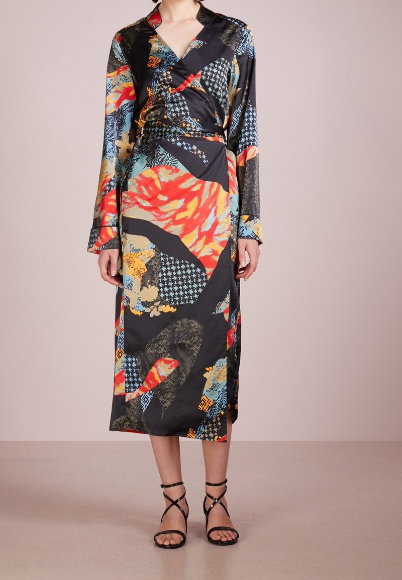 Femme portant une robe portefeuille à manches longues avec un imprimé abstrait coloré, accompagnée de sandales noires à talons hauts à lanières, sur un fond uni.
