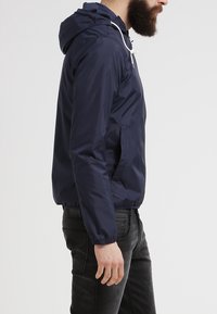 Marinblå vindjacka gjord av lättviktsmaterial, med huva, elastiska ärmmuddar och sidofickor. Slät, vattenavvisande yta.