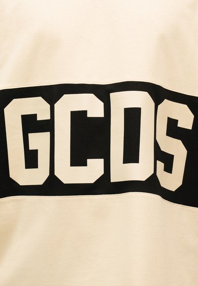 Μπλοκ μαύρου υφάσματος σε εκρού φόντο με μεγάλα λευκά γράμματα «GCDS» σε έντονη, γωνιακή γραμματοσειρά στο κέντρο.
