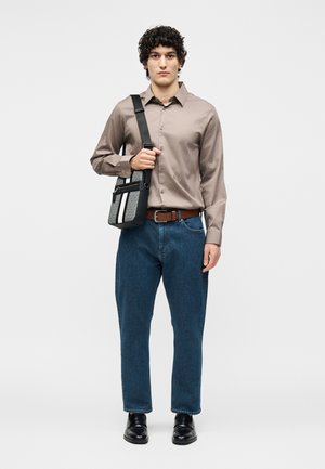 Joven de pie, vestido con una camisa beige abotonada, vaqueros azules, zapatos negros y sosteniendo una bolsa bandolera negra y gris con una franja blanca.