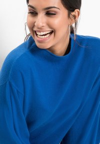 Maglione blu lavorato a maglia con collo alto e vestibilità ampia. Presenta una texture a coste e spalle a caduta. Il modello indossa orecchini in argento di grande effetto.