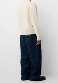 Personne portant un pull beige et un jean en denim bleu foncé à jambes larges avec de grandes poches arrière, debout devant un fond clair uni.