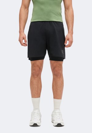 Mann trägt schwarze, geschichtete Sportshorts, weiße Sneakers, weiße Socken und ein grünes ärmelloses Top und steht vor einem schlichten Hintergrund.