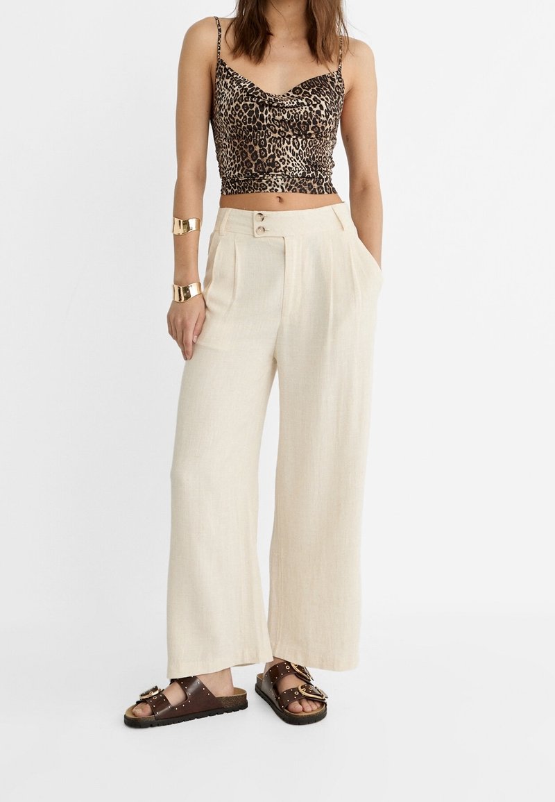 Femme portant un débardeur court imprimé léopard, un pantalon large crème taille haute, des bracelets manchettes dorés et des sandales marron cloutées.