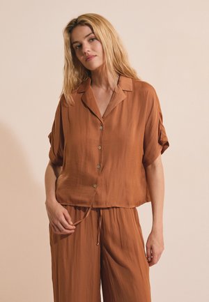 Donna con capelli biondi che indossa una camicia larga color ruggine con bottoni e pantaloni larghi abbinati su uno sfondo beige semplice.
