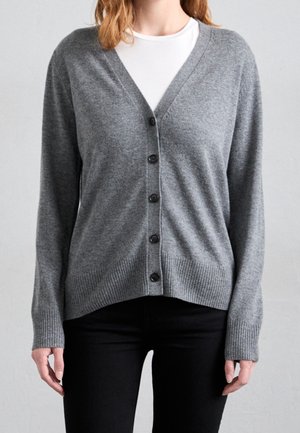 Frau trägt eine graue, geknöpfte Strickjacke über einem weißen Top mit schwarzen Hosen, steht vor einem schlichten hellen Hintergrund.
