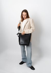 Femme en blazer beige, chemise blanche et jean bleu clair tenant un grand sac à bandoulière en cuir noir sur un fond blanc uni.