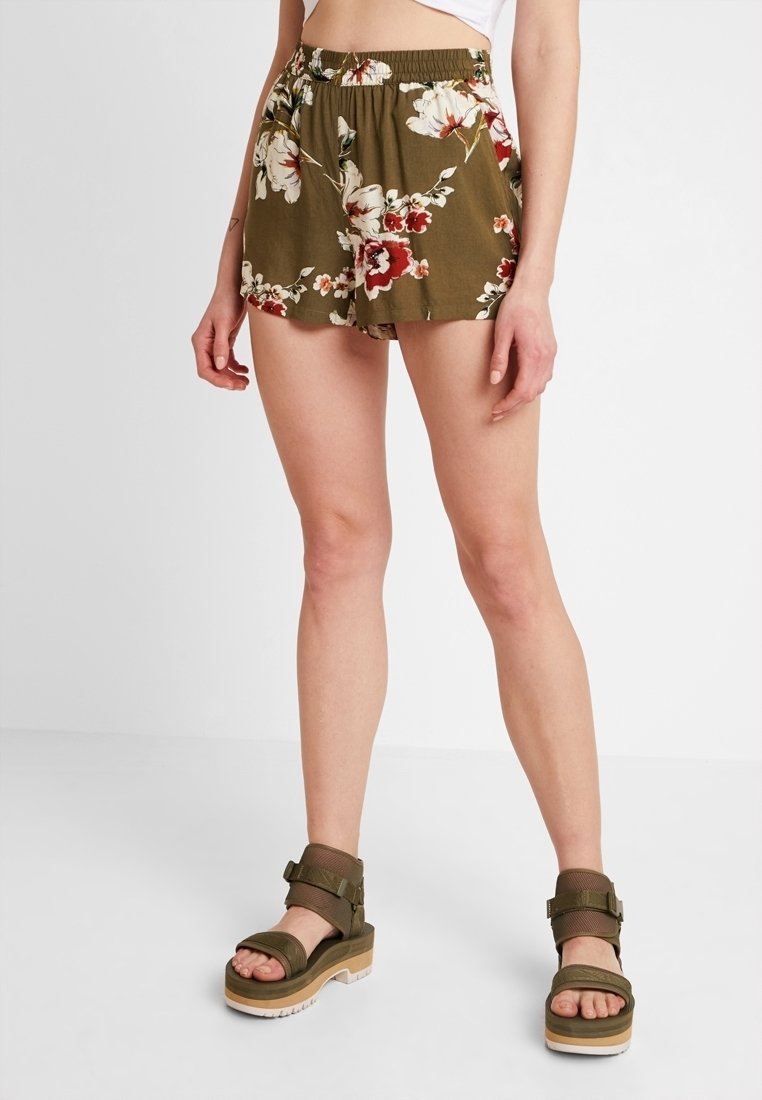 Shorts fleuris vert olive avec une taille froncée, ornés de fleurs rouges et blanches. Portés avec des sandales épaisses vert olive sur un fond blanc.