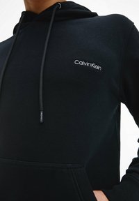 Felpa nera con cappuccio e cordini, tessuto morbido, tasca a marsupio e logo "Calvin Klein" ricamato bianco sul petto. Texture liscia.
