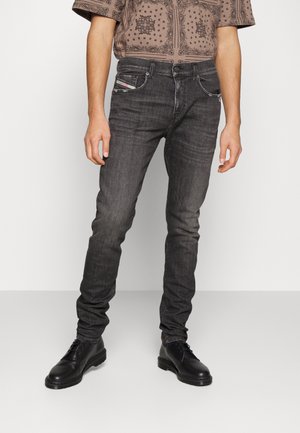 Homme portant un jean slim noir, des chaussures noires à lacets et une chemise marron à manches courtes avec motif, debout sur un fond blanc uni.