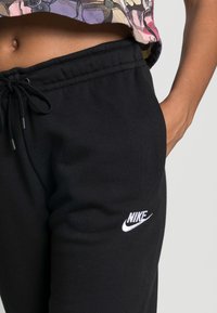 Černé tepláky vyrobené z měkké tkaniny mají stahovací pás a bílé logo Nike na pravé straně. Hladký povrch, uvolněný střih.