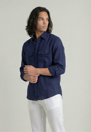 Man met haar tot op de schouders, gekleed in een donkerblauw overhemd met knopen en witte broek, kijkt naar links met gevouwen handen.