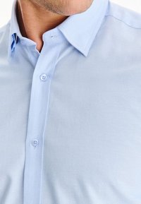 Mężczyzna w jasnoniebieskiej, zapinanej na guziki koszuli eleganckiej, z rozpiętym górnym guzikiem kołnierza, odsłaniającym część szyi i linii żuchwy.