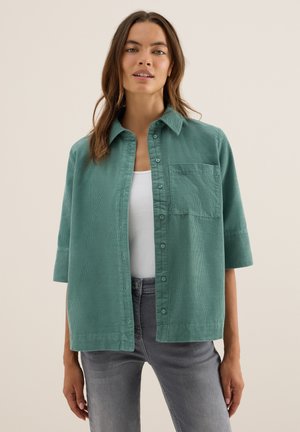Chemise verte à boutons en tissu texturé, à manches courtes, avec une poche poitrine, associée à un jean gris et un débardeur blanc.