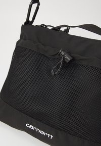 Musta laukku, jossa on verkkokankaasta valmistettu etutasku, päältä suljettava nyöri ja sileä kangaspohja. Logo näkyy valkoisena pohjassa.