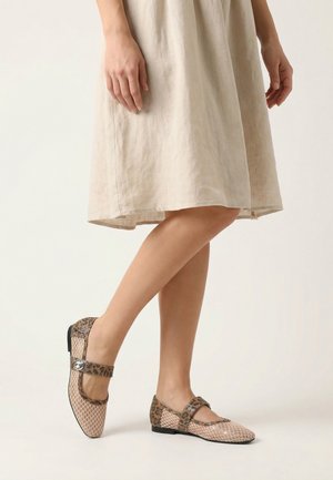 Kvinde iført beige kjole og beige spidse Mary Jane-flats med leopardprint stropper og lap nær hælen, stående på ét ben.