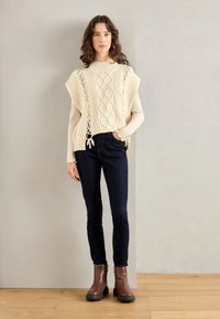 Gruby sweter z dzianiny w kolorze kremowym z detalami w splot kablowym, z bocznymi rozcięciami, zestawiony z ciemnymi skinny jeansami i brązowymi botkami za kostkę z teksturowanymi podeszwami.