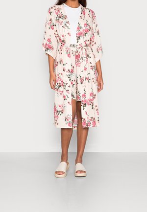 Kvinde iført en knælængde, cremefarvet kimono-robe med lyserøde blomster, bundet i taljen, hvid top under og beige sandaler med åben tå.