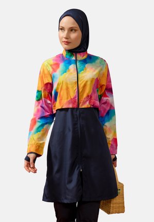 Kobieta nosząca kolorową krótka kurtkę tie-dye na granatowej sukience na zamek oraz granatowym hidżabie, trzymająca plecioną torebkę koszykową.
