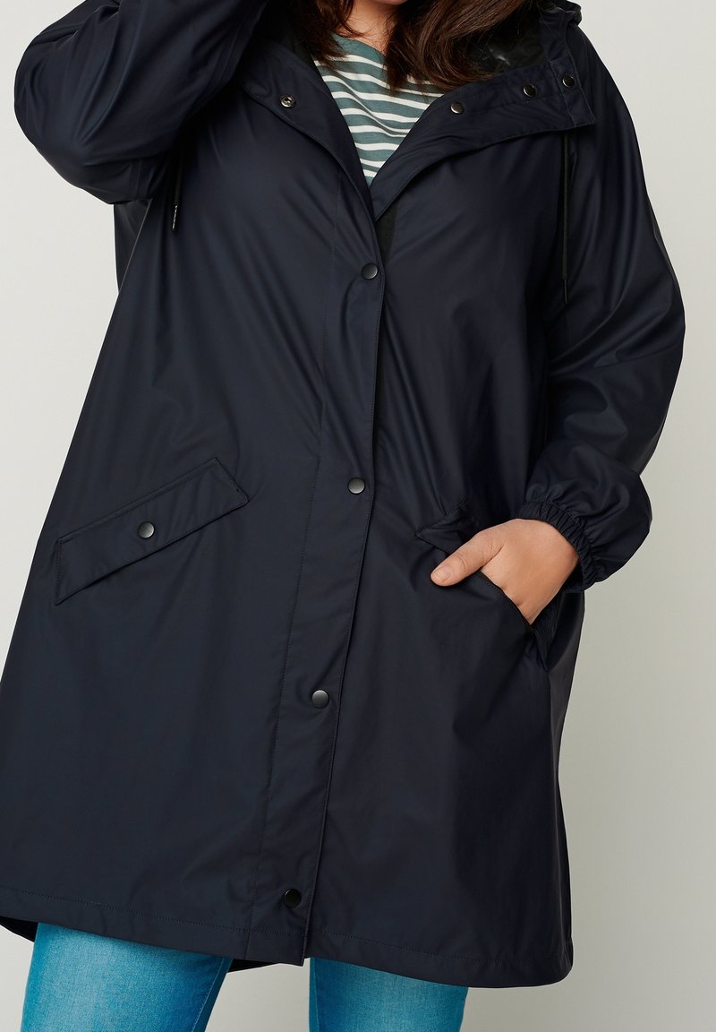 Personne portant un imperméable noir à capuche avec des boutons-pression, une main dans la poche, associé à un jean bleu et une chemise verte rayée en dessous.