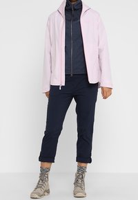 Světle růžová nepromokavá bunda s přiléhavým střihem, přes navy modrý fleecový zip. V kombinaci s tmavomodrými kalhotami a béžovými turistickými botami.