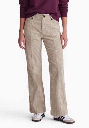 Pantalon en velours côtelé beige à jambes larges, fermeture triple bouton et zip, porté par une personne en baskets blanches et haut violet à manches longues.