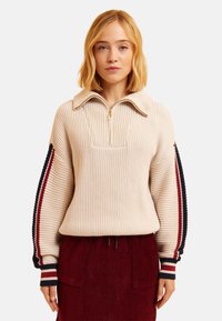 Pull en maille beige à col zippé, avec des accents rayés bleu marine et rouge sur les manches. Porté par-dessus une jupe en velours côtelé rouge foncé.