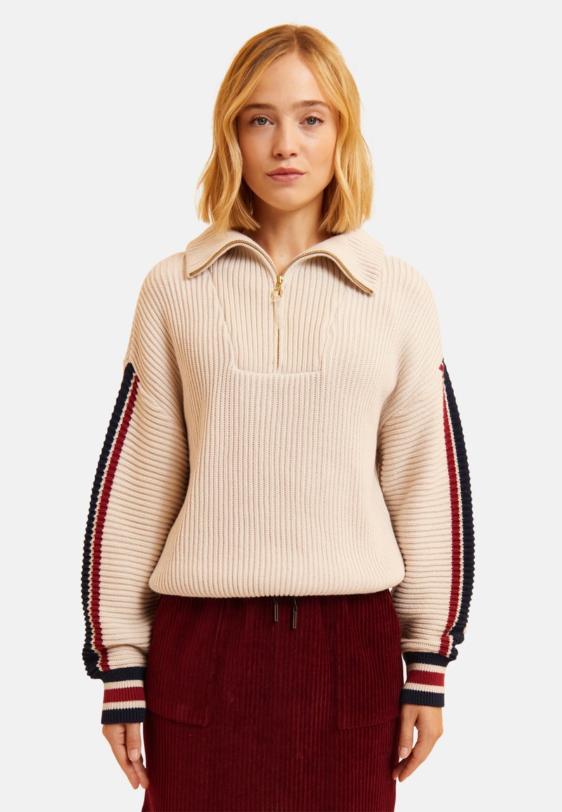 Pull en maille beige à col zippé, avec des accents rayés bleu marine et rouge sur les manches. Porté par-dessus une jupe en velours côtelé rouge foncé.
