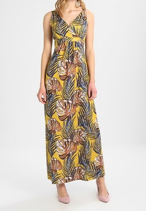 Woman wearing a sleeveless, yellow maxi dress with a tropical leaf pattern and pink high-heeled shoes, standing against a plain background.