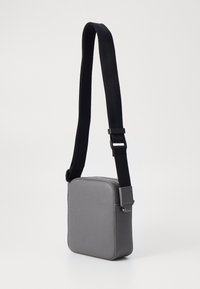 Borsa a tracolla sintetica grigia con una texture liscia, forma quadrata e una tracolla nera regolabile; presenta una chiusura a zip e un design minimalista.